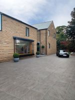 A large driveway in front of a modern brick house, laid with square Pietra Serena porcelain paving in a subtle mid-grey tone with a natural stone-effect finish.