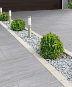 A sleek driveway or patio area featuring grey Vals porcelain paving with a subtle stone-effect texture, bordered by a decorative gravel bed with small green shrubs and bollard lighting.